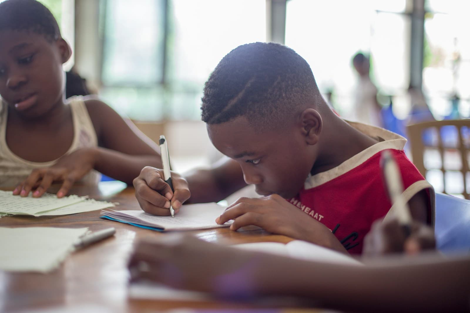 A boy in a red top leans in close, carefully writing in a small notebook at a shared table beside another child
