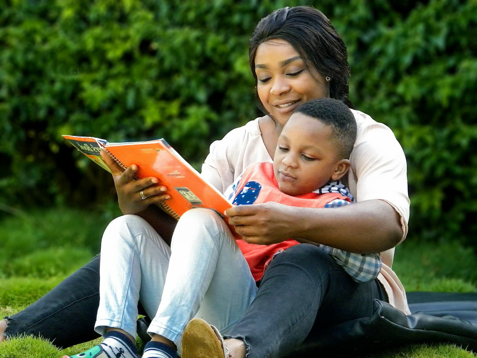 A woman sits on the grass with a young boy in her lap, reading an orange picture book together in front of a green hedge