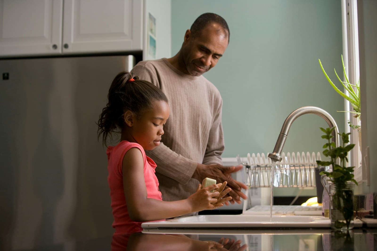 A father stands beside his young daughter at a kitchen sink, guiding her as she lathers soap and washes her hands