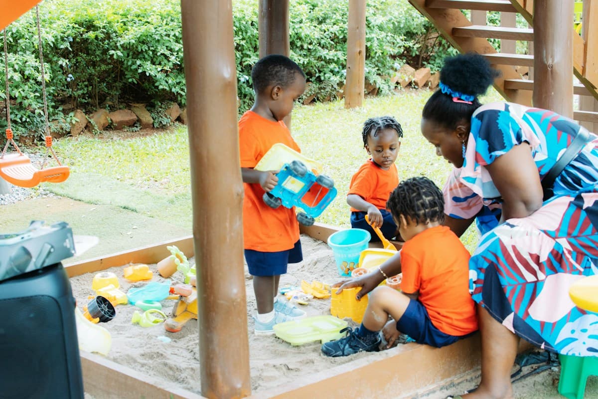 Young children playing with buckets and toy trucks in an outdoor sandbox while an educator joins in